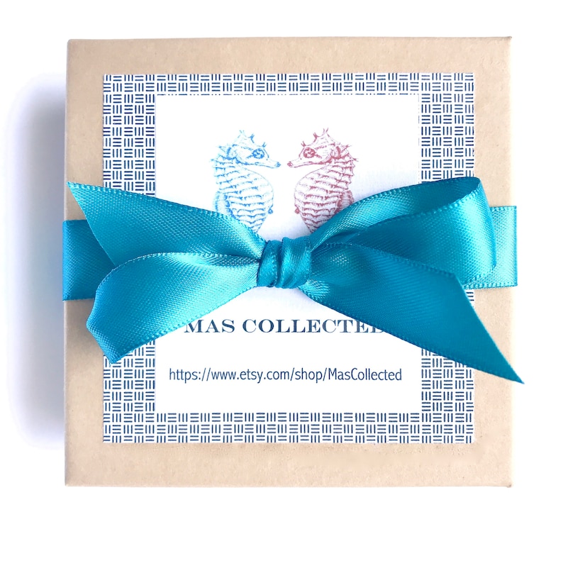 May include: A square, tan gift box tied with a bright teal satin ribbon. The box has a white label with two seahorse illustrations and the text "MAS COLLECTED" and a website address. The label is framed with a blue pattern.
