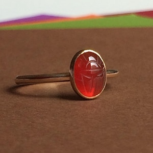May include: A gold ring featuring an oval-shaped, translucent orange-red carnelian gemstone. The stone is carved with a detailed scarab beetle design and is set in a smooth, polished gold bezel on a thin, plain gold band.