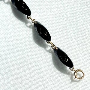 May include: A delicate necklace featuring black, oval-shaped beads linked by gold-colored chain. The necklace has a simple clasp closure. The beads have a glossy finish, reflecting light. The jewelry is set against a white background.