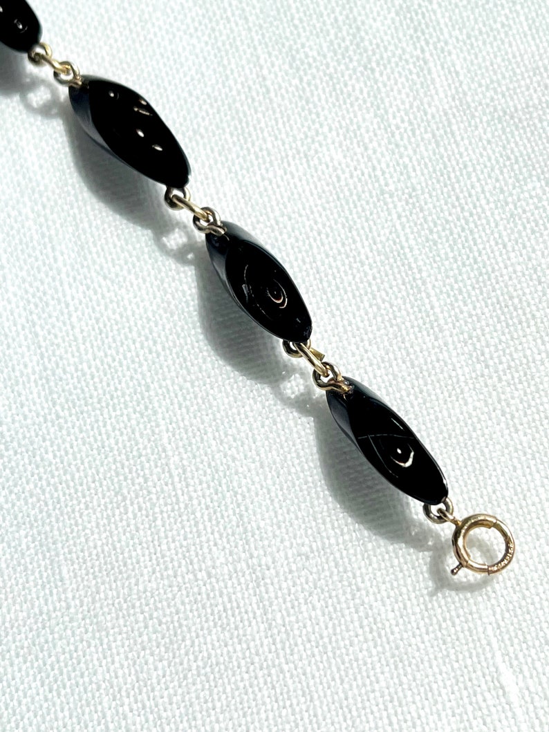 May include: A delicate necklace featuring black, oval-shaped beads linked by gold-colored chain. The necklace has a simple clasp closure. The beads have a glossy finish, reflecting light. The jewelry is set against a white background.