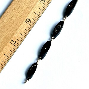 May include: A vintage black beaded necklace with oval-shaped beads connected by silver-tone metal links. The necklace is displayed on a white surface next to a wooden ruler.