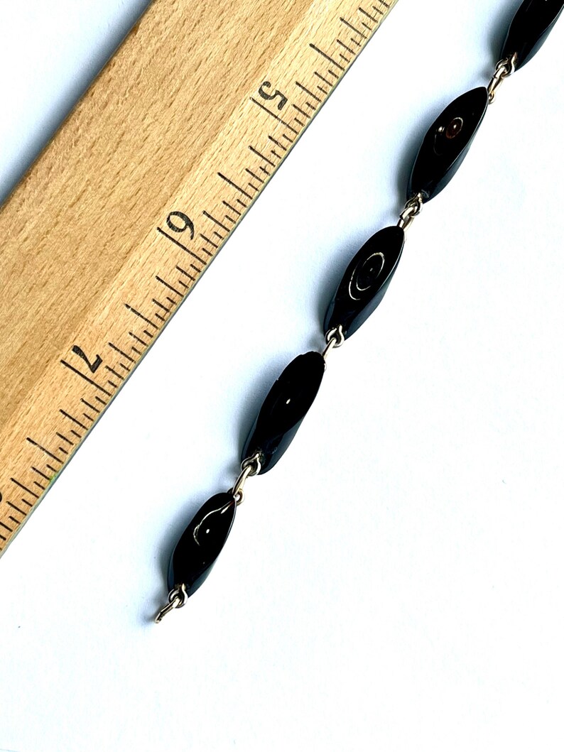 May include: A vintage black beaded necklace with oval-shaped beads connected by silver-tone metal links. The necklace is displayed on a white surface next to a wooden ruler.