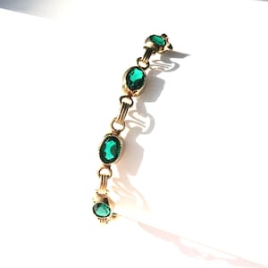 May include: A gold bracelet with five oval-shaped green gemstones. The bracelet is on a white background.