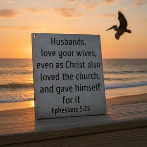 May include: A rectangular sign with the text "Husbands, love your wives, even as Christ also loved the church, and gave himself for it Ephesians 5:25" is displayed on a wooden surface. The background features a beach scene with a pelican in flight during sunset.