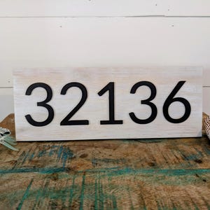 Może przedstawiać: Biały drewniany znak z czarnymi cyframi 32136. Znak znajduje się na drewnianej powierzchni.