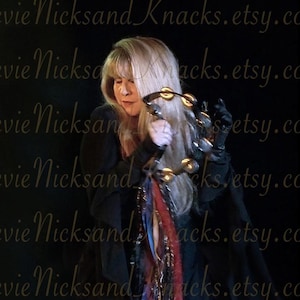 May include: A woman with long blonde hair is playing a tambourine. She is wearing a black dress with a long, flowing scarf. The background is dark.
