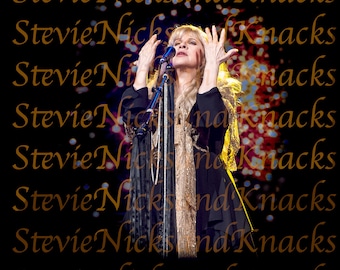 May include: Stevie Nicks performing on stage, wearing a black dress with a long, flowing scarf. The background is a dark, blurry image of a crowd and lights.