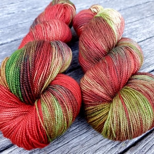 May include: Close-up of several skeins of hand-dyed yarn. The yarn is a mix of red, green, and brown colours. The yarn is arranged on a weathered wooden surface. The yarn is tightly wound and appears soft.