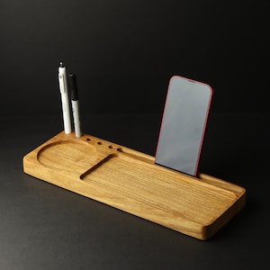 May include: A wooden desk organizer with a phone stand, two slots for pens, and a small tray. The organizer is made of light brown wood and has a natural finish.