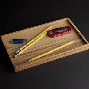 May include: A rectangular wooden tray with a natural wood grain finish. The tray holds yellow pencils, a red pocket knife, and a blue USB drive. The pencils have the text "Ticonderoga" printed on them.