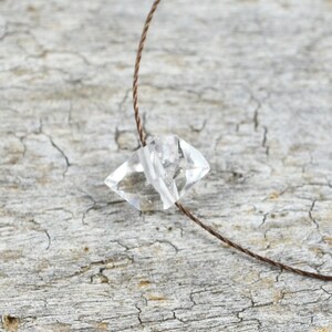 May include: A clear crystal pendant hangs from a thin brown cord. The pendant is a geometric shape with many facets. The crystal is resting on a piece of weathered wood.