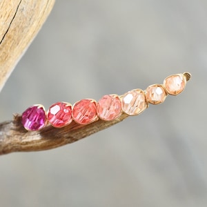 May include: A row of seven small, faceted gemstone beads in shades of pink, red, and orange, set in gold, arranged in a gradient on a wooden branch.
