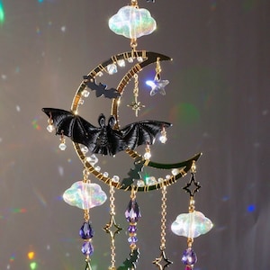 May include: A gold crescent moon wind chime with a black bat, iridescent clouds, and purple crystal beads. The chime features stars and bats hanging from the crescent moon.
