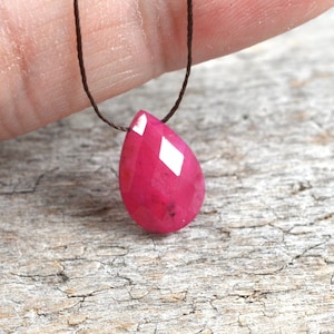 May include: A teardrop-shaped pink gemstone pendant on a brown cord necklace.