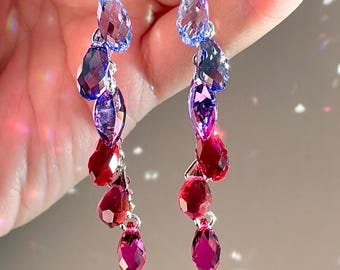 Puede incluir: Pendientes colgantes con un degradado de piedras preciosas de colores. Los pendientes presentan una serie de piedras facetadas en forma de lágrima en tonos azules, morados y rojos. Las piedras están suspendidas de ganchos plateados.