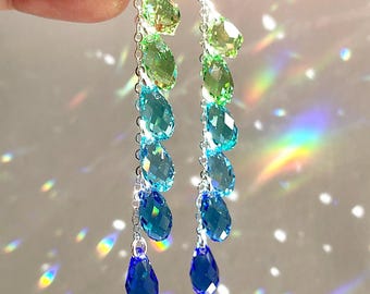 May include: A pair of drop earrings featuring a cascade of green, turquoise, and blue faceted crystals. The crystals are suspended from silver-tone hooks and chains. The earrings are displayed against a light background.