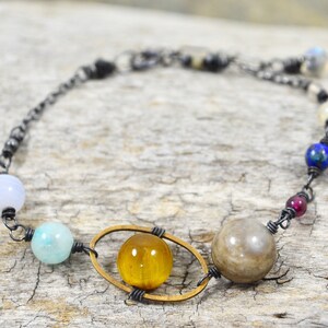 May include: A silver chain bracelet with a variety of colourful gemstone beads, including blue, green, brown, and yellow. The bracelet features a gold-toned oval frame with a tiger's eye bead inside.