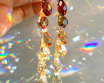 Puede incluir: Pendientes colgantes con una cascada de cristales facetados en forma de lágrima en tonos ámbar, marrón y transparente. Los pendientes están suspendidos de ganchos dorados. La luz atrapa los cristales, creando un efecto brillante.
