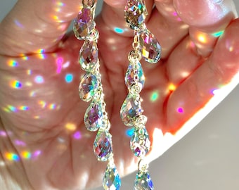 Könnte beinhalten: Hängende Ohrringe mit irisierenden, tropfenförmigen Kristallen. Die Ohrringe haben goldfarbene Haken und Verbindungselemente. Die Kristalle reflektieren einen Regenbogen an Farben und erzeugen einen funkelnden Effekt.
