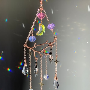May include: A delicate crystal and metal mobile with a crescent moon, stars, and teardrop-shaped crystals hanging from a branch-like structure. The mobile is made of rose gold metal and features iridescent crystals in shades of pink, purple, and blue.