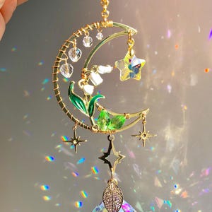 May include: A celestial-themed suncatcher featuring a gold-toned crescent moon, stars, and a large, faceted crystal ball. The moon design includes white flowers with green leaves and clear crystal beads. The suncatcher is approximately 10 inches in length.
