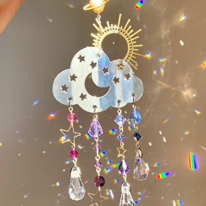 May include: A silver cloud-shaped suncatcher with a crescent moon and stars cut out. It has five strands of crystals hanging from it, each with a star charm and a teardrop-shaped crystal.
