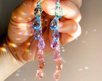 Puede incluir: Un par de pendientes colgantes con cristales en forma de lágrima en tonos azules, morados y rosas. Los pendientes son de plata y tienen un degradado de color.