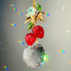 May include: A decorative crystal suncatcher with a gold chain, featuring red strawberry charms, gold leaves, and iridescent flowers. The suncatcher has a large, faceted crystal ball at the bottom. The design creates a rainbow effect.
