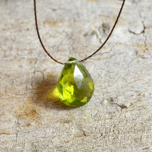 May include: A green gemstone pendant on a brown leather cord necklace. The pendant is a teardrop shape and has a faceted finish.