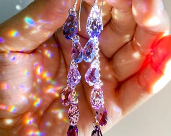 May include: A pair of long drop earrings featuring multiple faceted, teardrop-shaped crystals in a gradient of purple and violet hues. The crystals are linked together in a vertical chain and suspended from silver-toned metal earring hooks.