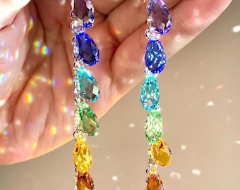 May include: A pair of long drop earrings featuring a vertical chain of seven faceted, teardrop-shaped crystals in a rainbow color gradient, starting with purple at the top and transitioning through blue, teal, green, yellow, orange, and red at the base.