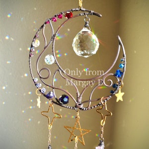 May include: A crescent moon-shaped wire sculpture with a crystal prism hanging from the center. The sculpture is adorned with small beads and crystals in shades of blue, purple, and pink.  The sculpture is suspended from a gold star and has three gold stars hanging from the bottom with amethyst crystals attached.