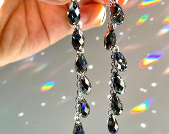 Könnte beinhalten: Ein Paar lange, baumelnde Ohrringe mit mehreren tropfenförmigen schwarzen Kristallen. Die Kristalle haben eine irisierende Oberfläche, die das Licht in mehreren Farben reflektiert.