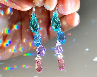 May include: A pair of drop earrings featuring a vertical arrangement of four faceted, teardrop-shaped crystals in a gradient of colours, transitioning from light blue at the top to dark blue, purple, and light pink at the bottom, suspended on silver hooks.