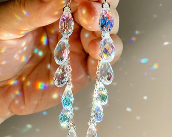 May include: A pair of long drop earrings featuring multiple faceted, clear crystal teardrop beads arranged in a vertical chain. The crystals exhibit a strong iridescent, rainbow-like light refraction effect. Each earring is attached to a silver-toned hook.