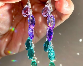 May include: A pair of dangling earrings featuring a vertical arrangement of faceted crystals in a gradient of colours, transitioning from light purple at the top to deep purple in the middle, and ending with teal and green crystals at the bottom.