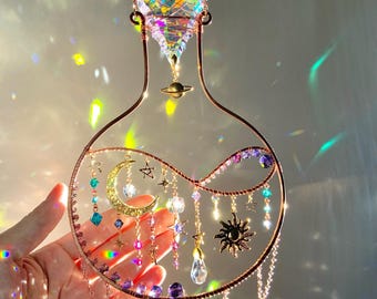Puede incluir: Un atrapasoles de temática celestial con un marco de alambre de color cobre. El diseño incluye un cristal facetado grande, una luna creciente, estrellas y colgantes de cristal. El atrapasoles está suspendido por una cadena.