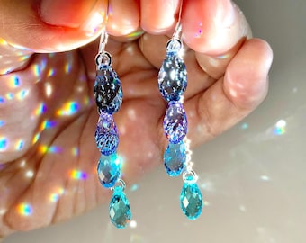 May include: A pair of drop earrings featuring four faceted, teardrop-shaped crystals in a gradient of blue shades, ranging from deep indigo at the top to bright turquoise at the bottom, suspended from silver-toned metal hooks.