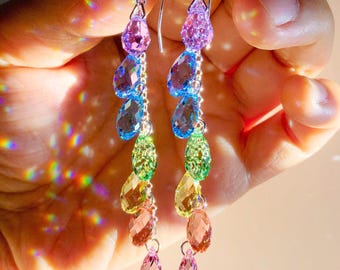 May include: A pair of long dangle earrings featuring a vertical chain of six faceted, teardrop-shaped crystals in a rainbow gradient of purple, blue, green, yellow, orange, and pink, suspended from silver-toned metal hooks.