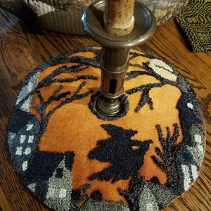 May include: A round orange and black Halloween-themed candle mat with a witch flying over a village. The mat is made of a textured fabric and has a candle holder in the center.