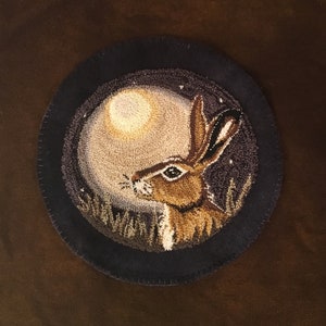 Puede incluir: Una alfombra redonda de color azul oscuro con un borde cosido presenta un conejo marrón mirando hacia arriba a una luna llena. La luna es de color beige claro y el conejo está rodeado de hierba verde.