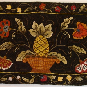May include: A hand-hooked rug with a dark brown background featuring a pineapple in a basket surrounded by stylized flowers and leaves. The rug has a border of vines and flowers.