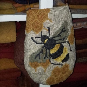 May include: A beige and brown wool rug with a bee design. The bee is black, yellow, and gray. The rug is hanging on a white shelf.