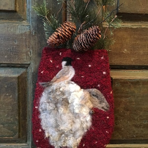 May include: A red felt stocking with a white fluffy bird design and a small bell. The stocking is decorated with white dots and is hanging on a wooden door with pine branches and pine cones.