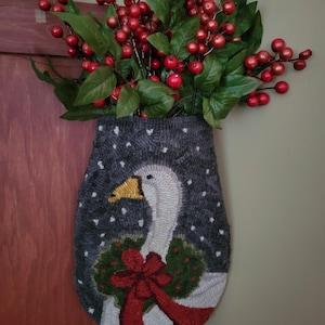 May include: A decorative wall hanging with a goose design, a wreath, and a red bow. The goose is white with a yellow beak, set against a grey background with white accents. The hanging is filled with red berries and green leaves.