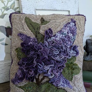 May include: A hand-stitched pillow featuring a large purple lilac flower with green leaves on a beige background.