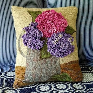 May include: A beige pillow with a hand-stitched design of a bouquet of purple and pink flowers in a green vase. The pillow is on a blue and white striped fabric.