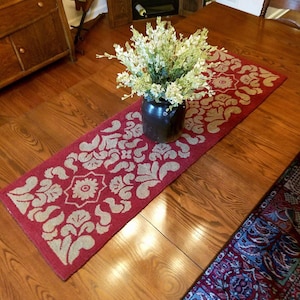 May include: A red table runner with a beige floral pattern, a bouquet of white flowers in a brown ceramic vase, and a wooden table.