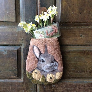 May include: A brown and tan needlepoint wall hanging featuring a rabbit with a white nose and a floral design around the rabbit. The wall hanging is hanging on a wooden door with a bouquet of white flowers in front of it.