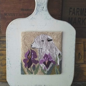 May include: A white sheep with a brown nose is looking at a purple iris flower. The sheep and flower are embroidered on a beige background.